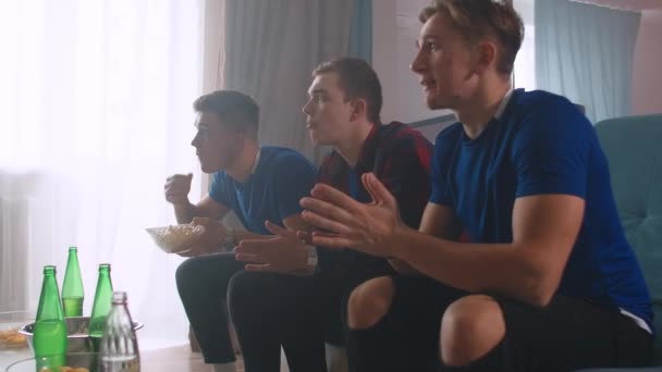 Three men in blue t shirts Watch a football match with friends sitting on the sofa and cheer and applaud looking directly at the camera - Footage, Video