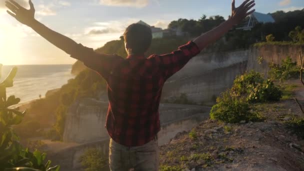 Young man traveler raising his hands high on top of the mountain above beautiful landscape on golden sunset - Video, Çekim