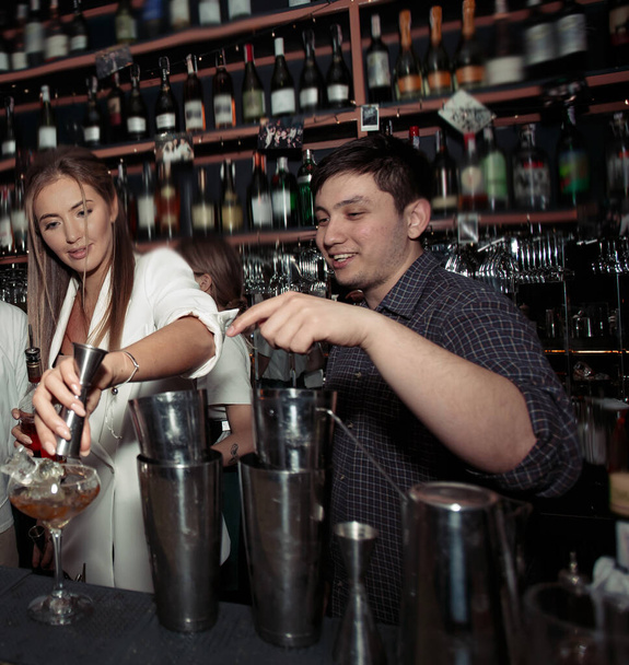 Profesjonalny barman, mężczyzna w szarej koszuli pokazuje młodej damie, jak zrobić smaczne koktajle i udekorować drinki. Butelki i szklanki stojące przy barze. Kobieta w białej nowoczesnej kurtce  - Zdjęcie, obraz