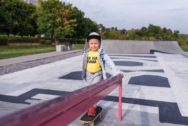 little boy 4-5 years old, rides on a skateboard, in summer in city on sports ground. Casual wear. Free space for copy text. Emotions of positive and fun, smile and joy on face. - Foto, afbeelding