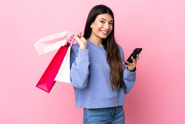 Young brunette girl over isolated pink background holding shopping bags and writing a message with her cell phone to a friend - Photo, Image