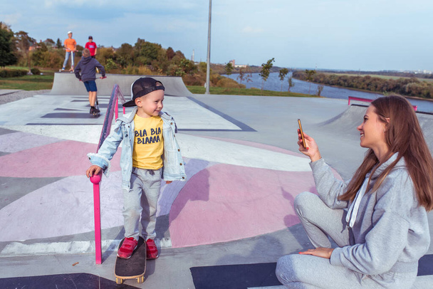 Young family, mother woman photographs son of a boy on phone, in summer in city. Relax and ride on a skateboard, positive emotions of joy and fun smile. Casual wear. - 写真・画像