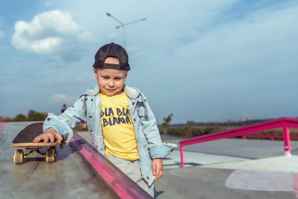 little boy 4-5 years old, rolls a skateboard, in summer in city on sports field sits. Casual wear denim and baseball cap. Free space for copy text. Emotions of positive and joy smile. - Fotoğraf, Görsel