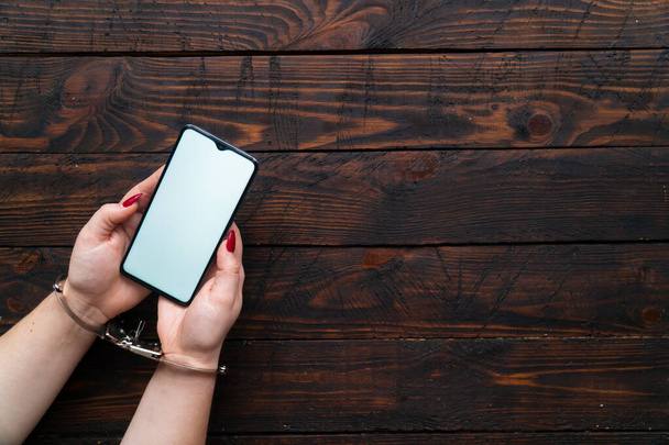 Κοντινό πλάνο γυναίκα χέρια σε χειροπέδες κρατήσει ένα smartphone με μια κενή οθόνη για κείμενο ή το λογότυπο. Η έννοια της διαδικτυακής εξάρτησης, της κοινωνικής δικτύωσης και των gadgets. - Φωτογραφία, εικόνα