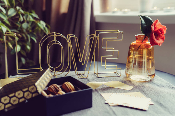 Festive box with chocolate macaroons cakes, rose flower in vase, postcard and wire word Love on gray napkin. Cozy gift idea for lover. Hygge Valentine's day concept. Romantic mood in noir nordic style. - Photo, Image
