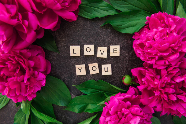 Close up Love you text on wooden blocks border with the frame made of fresh pink peonies with green leaves on black stone background. Flat lay, top view. Floral valentine's day card - Фото, изображение