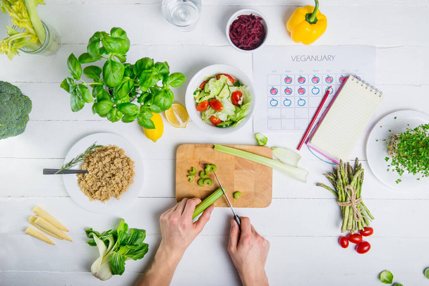 Мужчина режет сельдерей и готовит веганский свежий салат с овощами на ужин. Вегетарианский календарь и ежедневное планирование питания. Мужские руки закрыть вид сверху, вегетарианские ингредиенты и на белом фоне
 - Фото, изображение