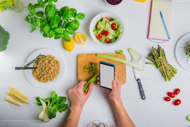 上からの眺め白い画面のスマートフォンを手に持ち、まな板の上にセロリの棒を持つ男の手。野菜とビーガン新鮮なサラダを調理.ダイエット計画。持続可能な植物性食品 - 写真・画像