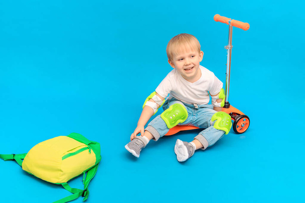 Een jongetje zit op een scooter ter bescherming tegen schaafwonden en blauwe plekken, kniebeschermers en elleboogstukken en lacht. Studio fotografie met plaats voor tekst op een blauwe achtergrond. - Foto, afbeelding