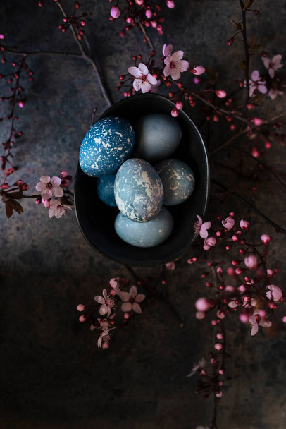 Blue Easter Eggs on black bowl, hand painted with natural dyes, surrounded by pink almond blossoms on rustic surface. overhead view - Foto, Imagen