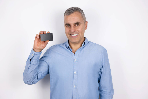 adult handsome smiling man with grey hair wearing blue shirt holds bank card looks happy standing on isolated white background. - Foto, Imagen