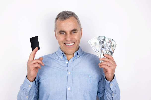 adult man with grey hair wearing blue shirt holds bank card in one hand and cash in another looks happy standing on isolated white background. - Фото, изображение