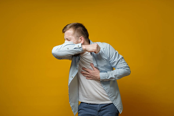 Mężczyzna w masce medycznej kaszle w łokciu i trzyma rękę na klatce piersiowej, doświadczając bólu. Koncepcja rozprzestrzeniania się wirusa. - Zdjęcie, obraz