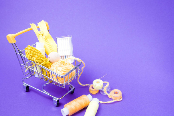 shopping trolley with various accessories and accessories for sewing, yellow threads of different colors, sewing machine needles, ball of thread on bright purple background copy space, hobby shopping concept - Fotoğraf, Görsel