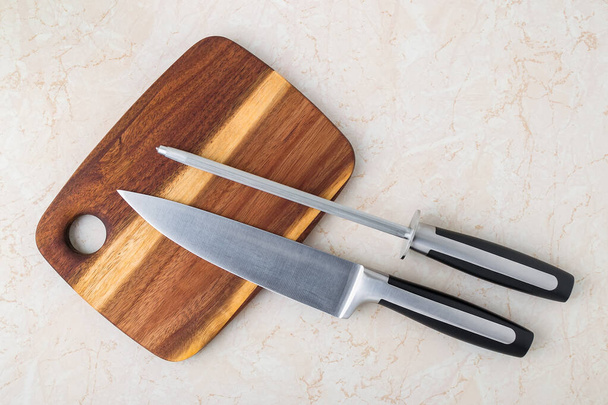 Professional chef knife and a sharpening steel on a wooden cutting board over a kitchen table. Modern kitchen utensils made of high carbon molybdenum vanadium steel. Top view.  - Foto, Imagem