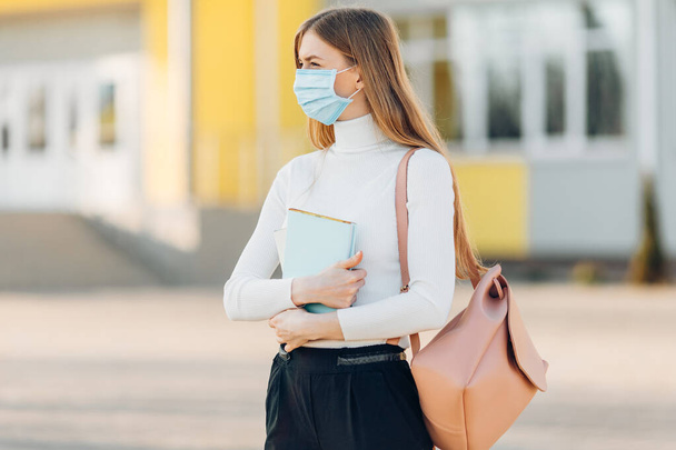 A young girl in the background of a building wears a face mask that protects against the spread of coronavirus disease. Close- up of a young woman with a surgical mask on her face against SARS-cov-2. - Photo, image