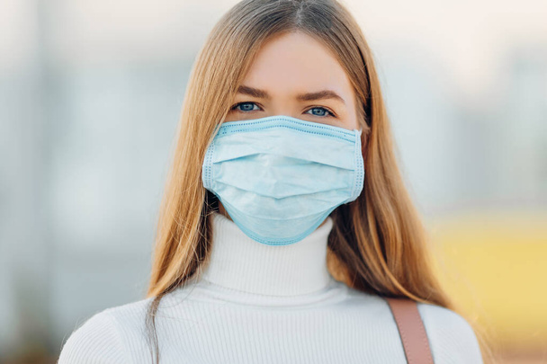 A young girl in the background of a building wears a face mask that protects against the spread of coronavirus disease. Close- up of a young woman with a surgical mask on her face against SARS-cov-2. - Φωτογραφία, εικόνα