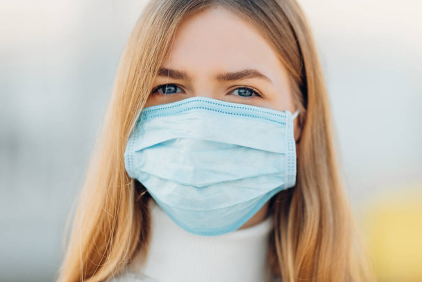 A young girl in the background of a building wears a face mask that protects against the spread of coronavirus disease. Close- up of a young woman with a surgical mask on her face against SARS-cov-2. - Foto, Bild