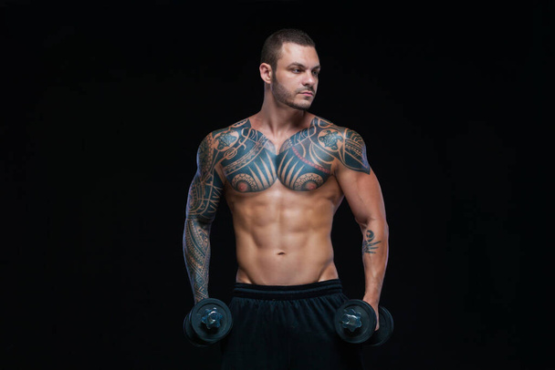 Tätowierter kräftiger muskulöser, athletischer Mann, der mit Kurzhanteln auf schwarzem Hintergrund Muskeln aufpumpt - Foto, Bild