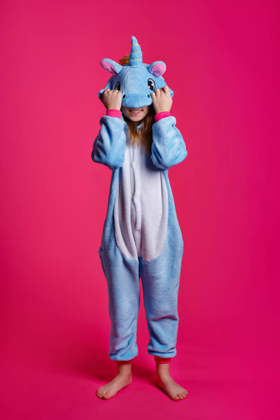 Charming girl playfully poses in the costume of the unicorn. Studio shot of an emotional woman in kigurumi having fun on a pink background. - Photo, Image