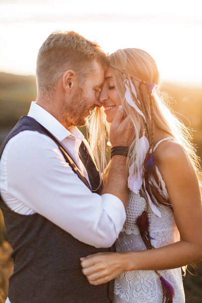 beautiful couple dressed in boho style embracing each other in the sunny summer field. Handsome man in casual wear and woman in white dress and feathers in hair walking in the field at sunset - Fotó, kép