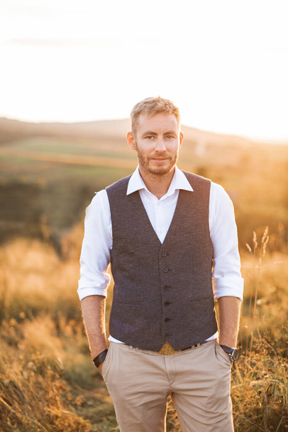 Portrait of handsome retro stylish man, posing on the background of summer field, sunset. Young bearded blond man wearing shirt, vest and pants, standing outdoors in the summer field - Фото, зображення
