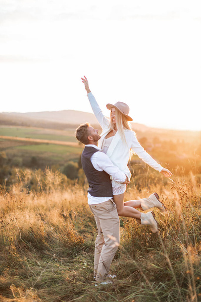 Shot of young boho hippie woman being carried by her handsome boyfriend in summer field. Couple having fun on their summer evening outdoors - Zdjęcie, obraz