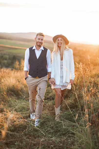 Handsome stylish man in shirt, vest and pants and pretty boho woman in dress, jacket and hat walking in the field with straw bales, holding hands and smiling. Summer evening, sunset - Foto, afbeelding