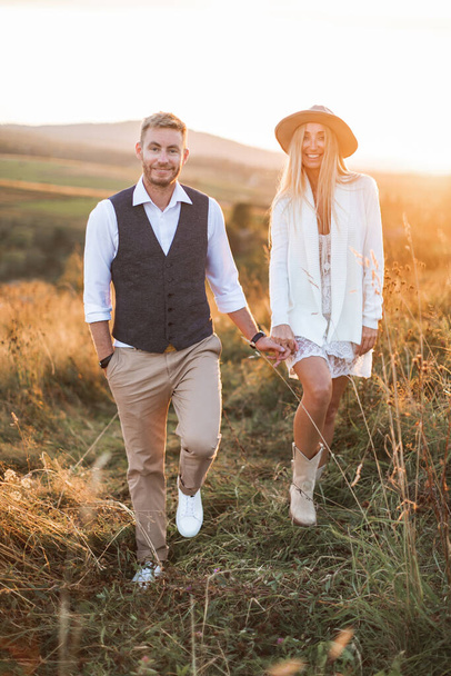 summer, love, romance and people concept - happy smiling young hippie cowboy style couple holding hands and walking in summer field, outdoors. Woman in dress and cowboy boots, man in casual suit - Foto, afbeelding