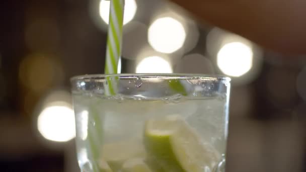 Close up glass of sparkling water with mint, lime and ice on a blurry lights background. Barman stirring cocktail. Jumping water droplets are clearly visible in backlit. - Footage, Video