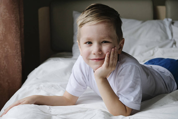 Lindo niño pequeño acostado en la cama blanca, mirando a la cámara y sonriendo. Concepto de estilo de vida
 - Foto, imagen