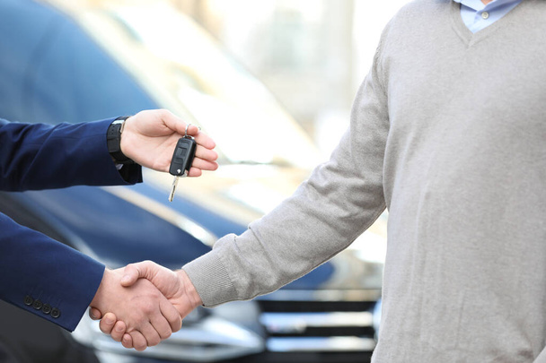 Salesman giving key to customer while shaking hands in modern auto dealership, closeup. Buying new car - Zdjęcie, obraz