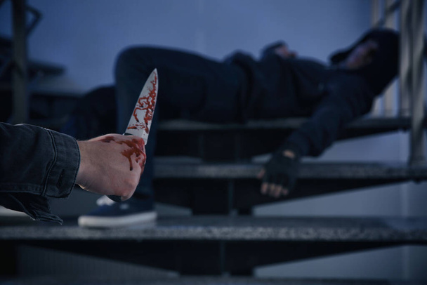 Man with bloody knife and unconscious person on stairs indoors, space for text. Dangerous criminal - Foto, immagini