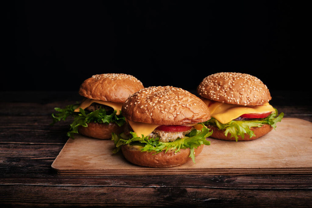 肉、チーズ、レタス、トマトを木の板に乗せた3つのジューシーなハンバーガー - 写真・画像