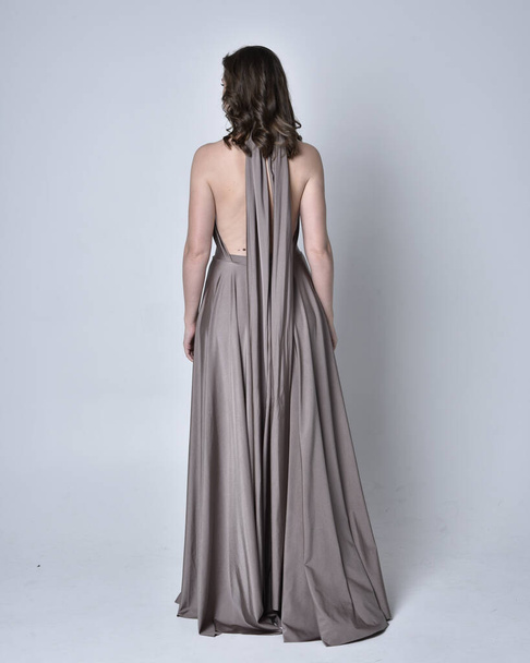 Portrait of a pretty brunette girl wearing a long silver evening gown, full length standing pose against a studio background - Foto, afbeelding