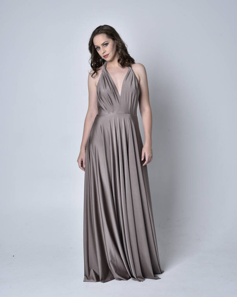 Portrait of a pretty brunette girl wearing a long silver evening gown, full length standing pose against a studio background - Foto, Bild