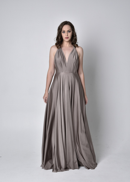 Portrait of a pretty brunette girl wearing a long silver evening gown, full length standing pose against a studio background - Foto, imagen