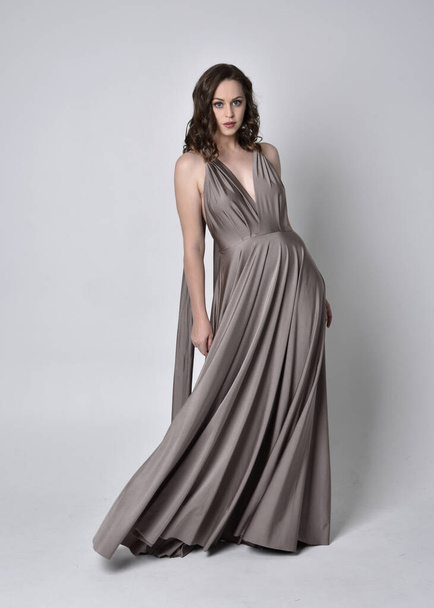 Portrait of a pretty brunette girl wearing a long silver evening gown, full length standing pose against a studio background - Foto, afbeelding