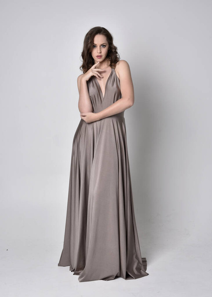 Portrait of a pretty brunette girl wearing a long silver evening gown, full length standing pose against a studio background - Photo, image