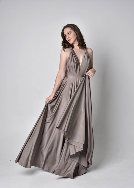 Portrait of a pretty brunette girl wearing a long silver evening gown, full length standing pose against a studio background - Φωτογραφία, εικόνα