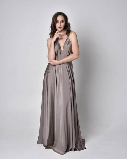Portrait of a pretty brunette girl wearing a long silver evening gown, full length standing pose against a studio background - Photo, Image