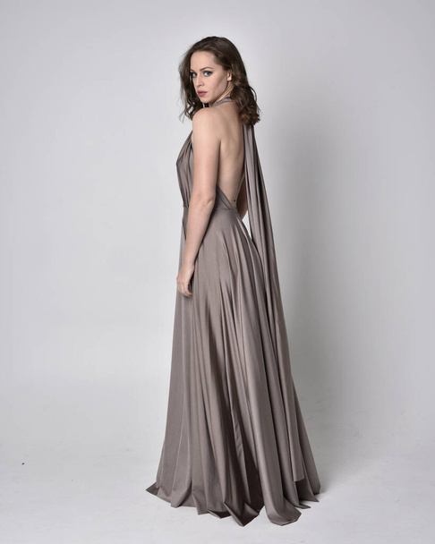 Portrait of a pretty brunette girl wearing a long silver evening gown, full length standing pose against a studio background - Photo, Image