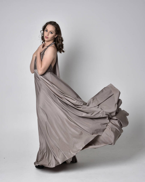 Portrait of a pretty brunette girl wearing a long silver evening gown, full length standing pose against a studio background - Foto, Imagem
