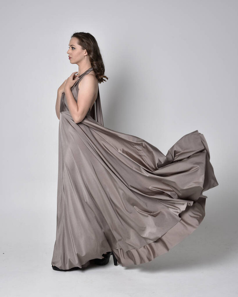 Portrait of a pretty brunette girl wearing a long silver evening gown, full length standing pose against a studio background - 写真・画像