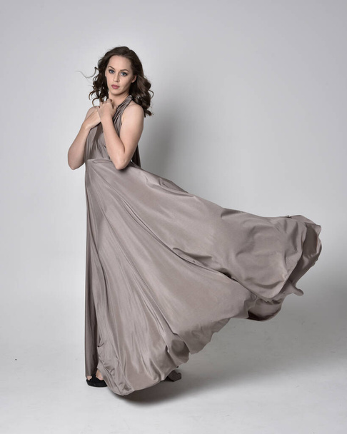 Portrait of a pretty brunette girl wearing a long silver evening gown, full length standing pose against a studio background - Foto, immagini