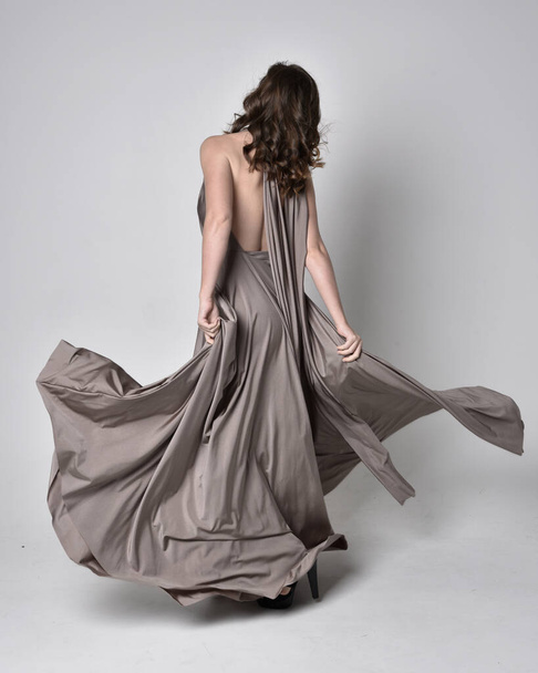 Portrait of a pretty brunette girl wearing a long silver evening gown, full length standing pose against a studio background - Foto, Imagen
