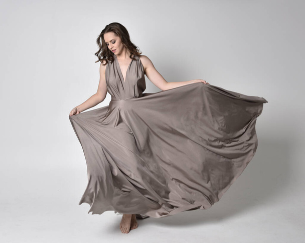 Portrait of a pretty brunette girl wearing a long silver evening gown, full length standing pose against a studio background - Foto, Imagem