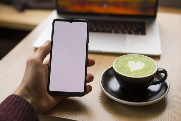 Lugar de trabajo con café con leche matcha, teléfono inteligente de mano masculino con pantalla en blanco
 - Foto, Imagen