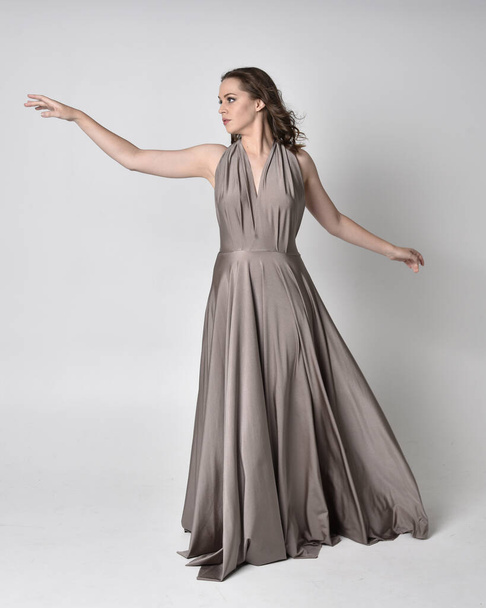 Portrait of a pretty brunette girl wearing a long silver evening gown, full length standing pose against a studio background - Fotografie, Obrázek