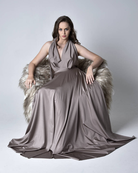 Retrato de uma menina bonita morena vestindo um longo vestido de noite de prata, comprimento total sentado pose na cadeira contra um fundo de estúdio
. - Foto, Imagem